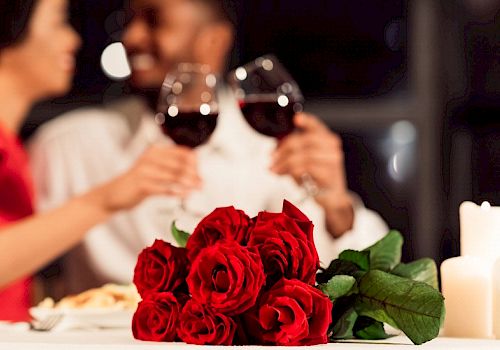 A romantic dinner setup with a bouquet of red roses, two people toasting with wine glasses, and candles on a softly lit table.