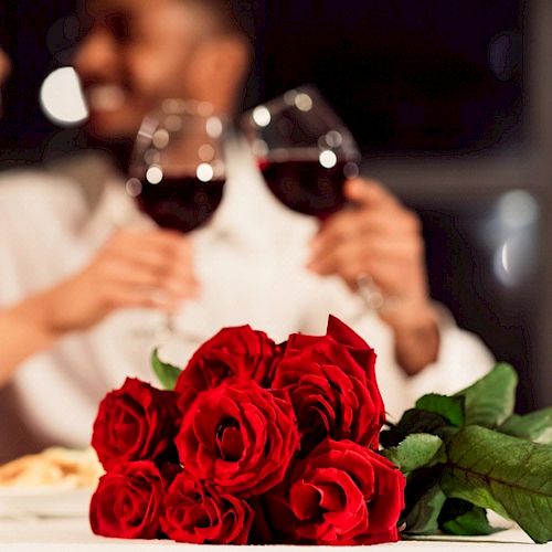A romantic dinner setup with a bouquet of red roses, two people toasting with wine glasses, and candles on a softly lit table.