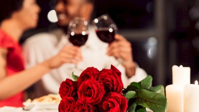 A romantic dinner setup with a bouquet of red roses, two people toasting with wine glasses, and candles on a softly lit table.