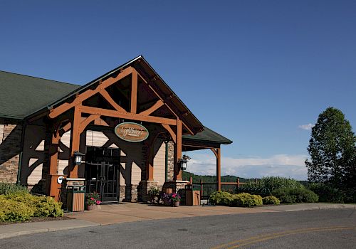 The image features a rustic building with wooden beams, a sign above the entrance, and landscaped greenery under a clear blue sky.