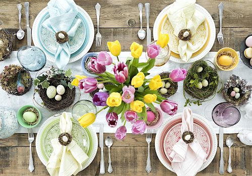 A beautifully decorated spring-themed table setting with colorful plates, napkins, tulips, nests, eggs, and various tableware, arranged atop a wooden table.