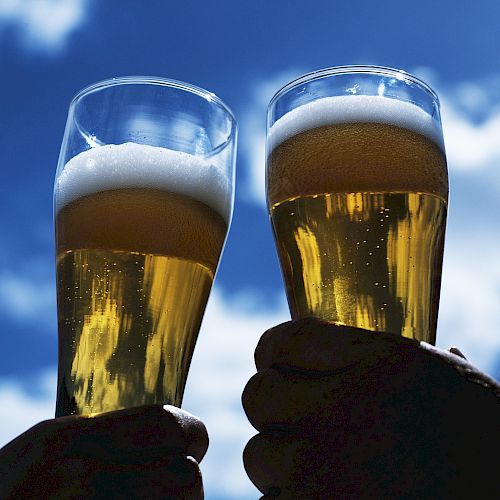 Two hands holding up glasses of beer against a bright blue sky with fluffy clouds in the background.