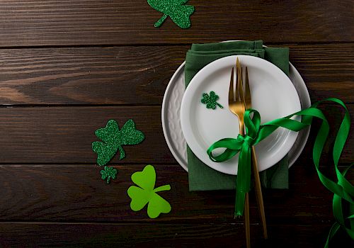 A festive table setting with a white plate, green napkin, gold utensils, green ribbon, and shamrock decorations on a wooden surface.
