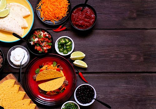 The image shows a variety of ingredients for making tacos, including tortillas, shredded cheese, salsa, sour cream, diced vegetables, and lime wedges.