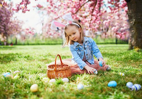 A child wearing bunny ears collects colorful Easter eggs in a grassy, flower-filled park with a woven basket beside her.