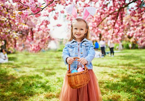 A young girl with bunny ears stands under pink blossom trees, holding an Easter basket. She smiles, dressed in a denim jacket and pink dress.