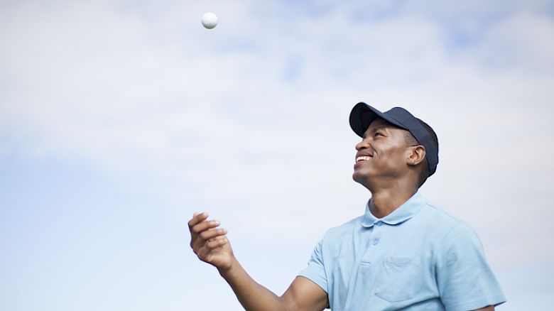A man wearing a blue polo shirt and cap is smiling while tossing a ball in the air, set against a cloudy sky background.