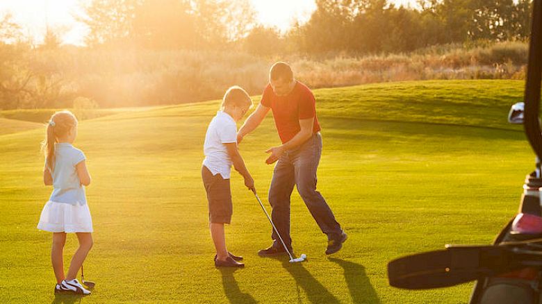 An adult is teaching a child how to putt on a golf course while another child looks on in the background, during a sunny day.