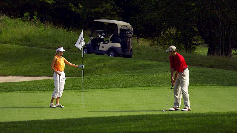 Two golfers stand on a green, one holding a flagstick while the other lines up a putt. A golf cart is parked in the background near the trees.