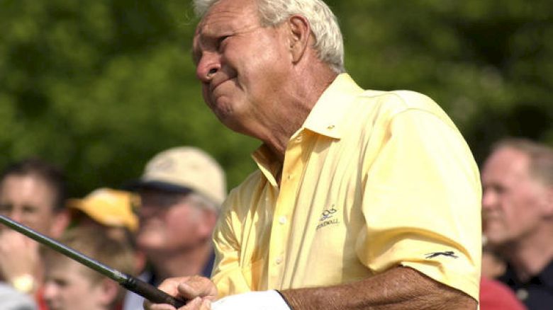 An older man in a yellow shirt swings a golf club, surrounded by onlookers with a green background in the outdoors, appearing focused.