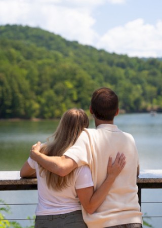 A couple embraces while looking out at a scenic river surrounded by lush green hills in a peaceful outdoor setting.