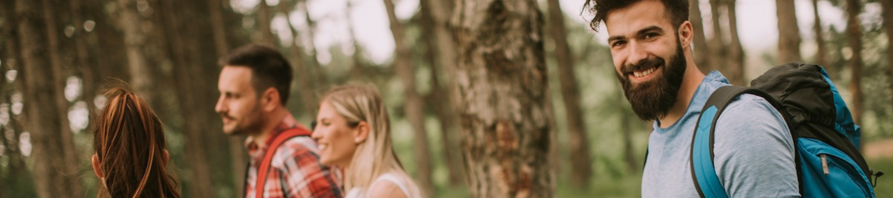 Four people are walking through a forest; they appear to be hiking, with one person smiling at the camera and carrying a backpack, ending the sentence.