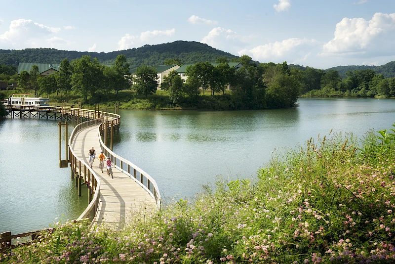 The image depicts a scenic, curved boardwalk bridge over a body of water, with people walking on it, surrounded by greenery and hills in the background.