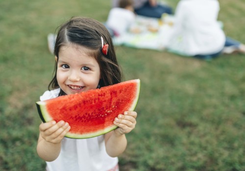 A child is holding a slice of watermelon and smiling, with people having a picnic in the background on a grassy area.