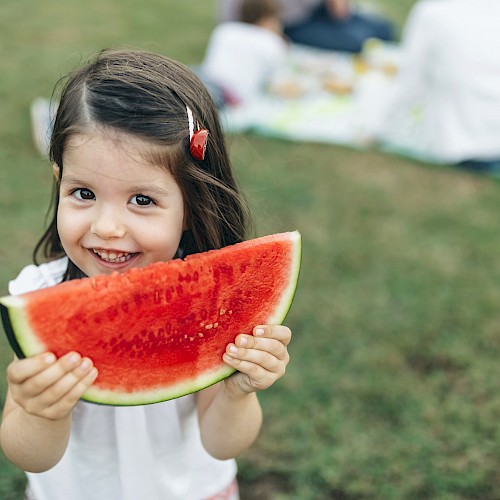 A child is holding a slice of watermelon and smiling, with people having a picnic in the background on a grassy area.