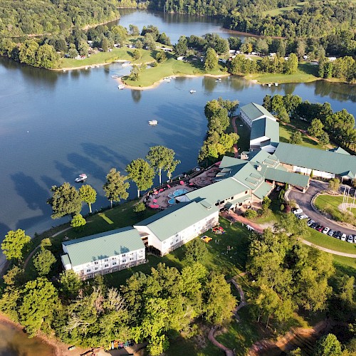 An aerial view of a large building complex surrounded by lush greenery and water bodies, with boats visible on the water and a parking area.