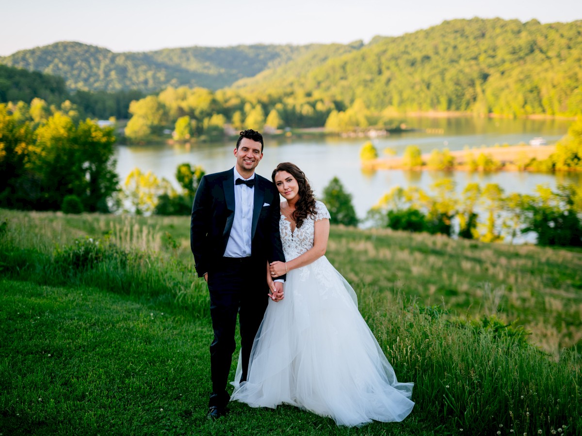 A couple in wedding attire stands on a grassy field with a lake and forested hills in the background, smiling and holding hands.
