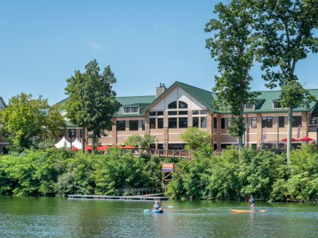 A large building with green roofs stands by a riverside, surrounded by trees, with people kayaking on the calm water.