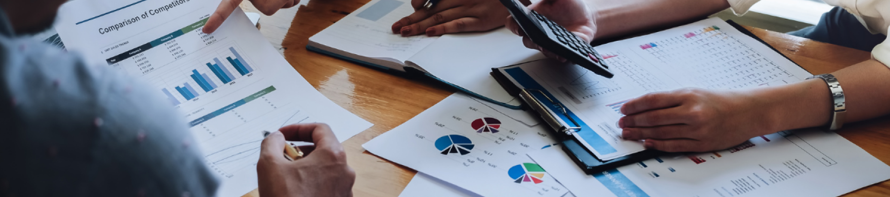 Three people are in a meeting, analyzing graphs and charts on paper documents and a laptop. One person is holding a calculator.