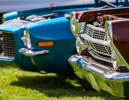 The image shows the front ends of two classic cars, one blue and one maroon, parked on grass.