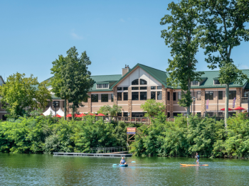 The image shows a waterside building surrounded by trees, with two people kayaking on the water in the foreground, under a clear sky.