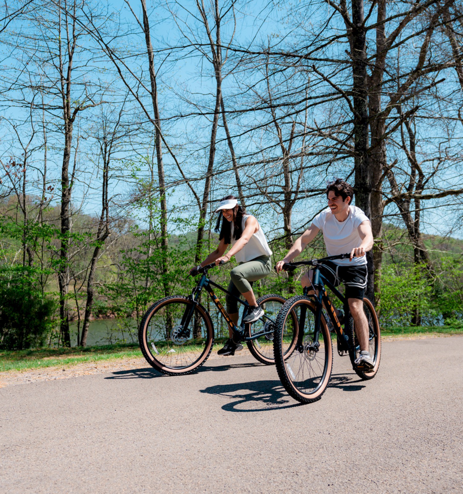 Two people are riding bikes on a paved path in a scenic, wooded area on a sunny day with clear blue skies.