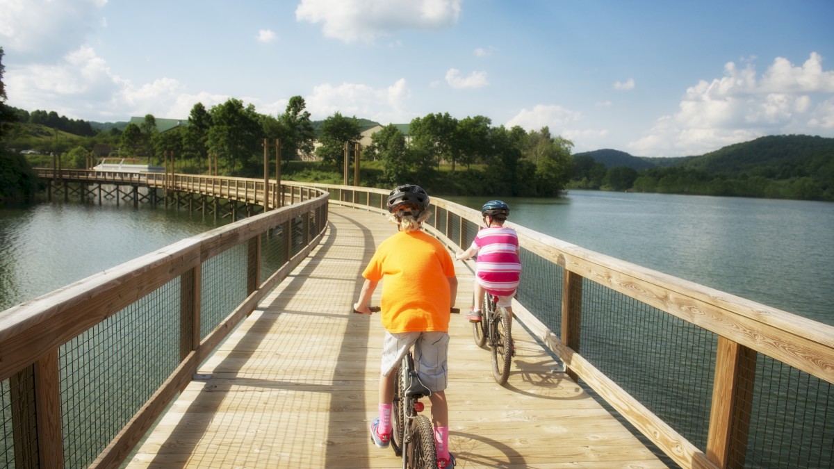 Two people are riding bicycles on a wooden boardwalk over a lake, surrounded by green trees and hills under a partly cloudy sky.