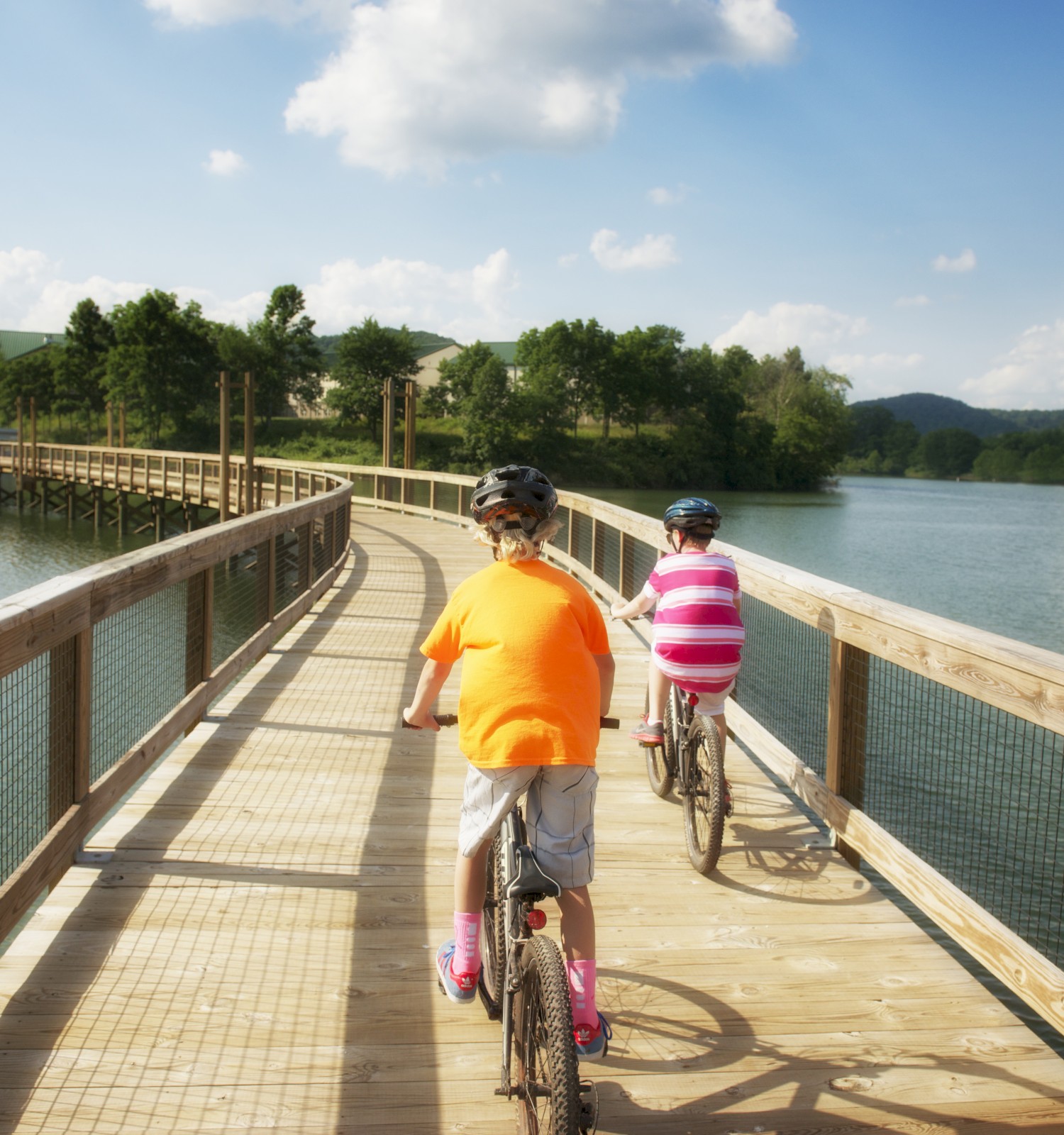 Two people are riding bicycles on a wooden boardwalk over a lake, surrounded by green trees and hills under a partly cloudy sky.