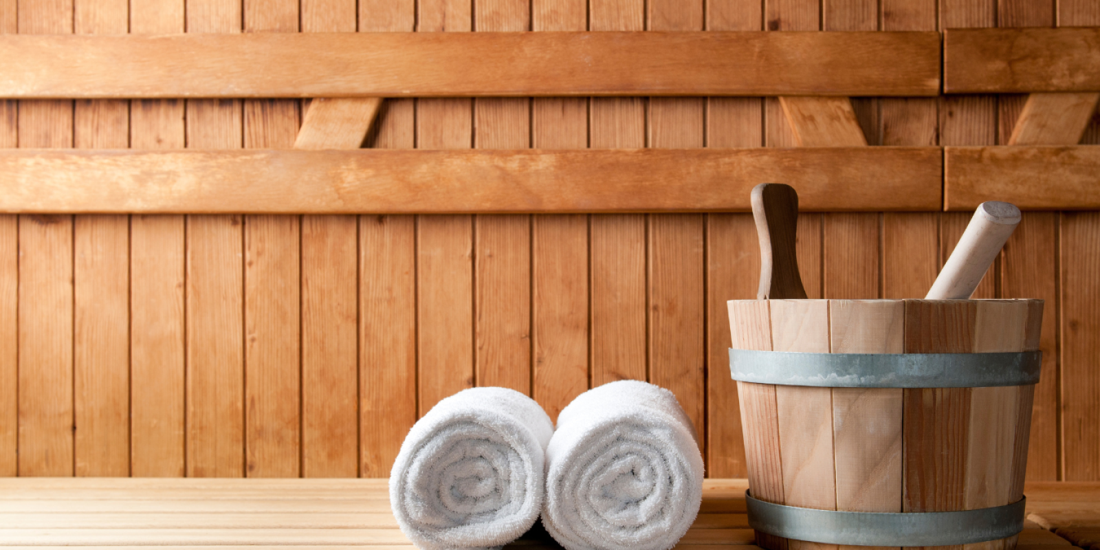 The image shows a sauna with wooden walls, a wooden bucket with a ladle, and two neatly rolled white towels placed on a bench.