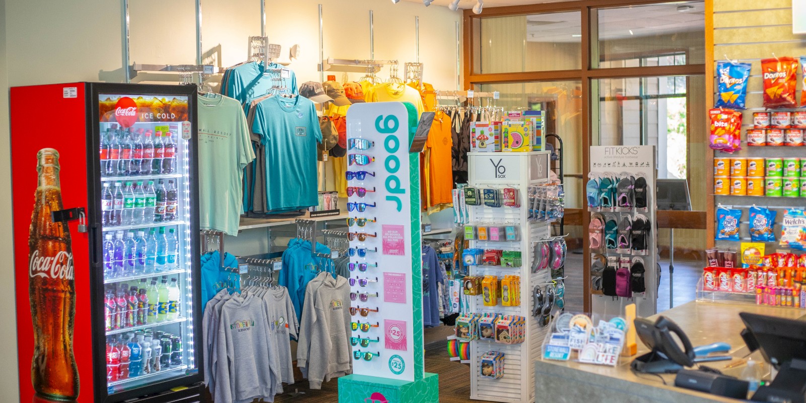 The image shows a convenience store with a Coca-Cola fridge, clothing on racks, sunglasses display, snacks, and a counter with a register.