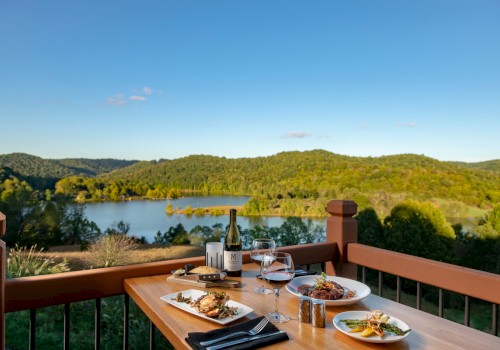 A table on a balcony with wine, glasses, and plates of food, overlooking a lake and forested hills under a clear blue sky.