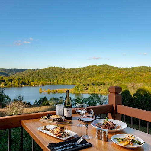 A table on a balcony with wine, glasses, and plates of food, overlooking a lake and forested hills under a clear blue sky.