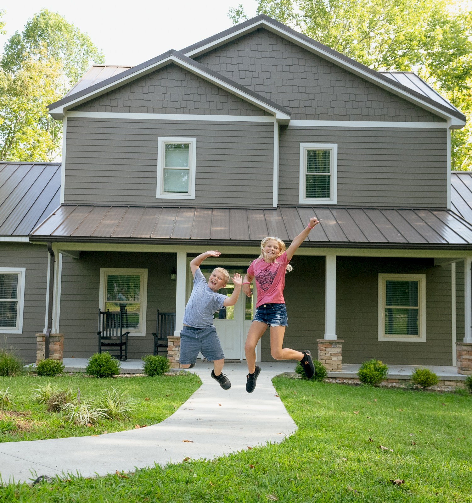 Two children are jumping with joy outside a modern, gray house surrounded by greenery on a sunny day.