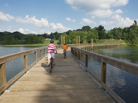 Two individuals are cycling on a wooden bridge over a body of water surrounded by lush greenery and trees, under a partly cloudy sky.