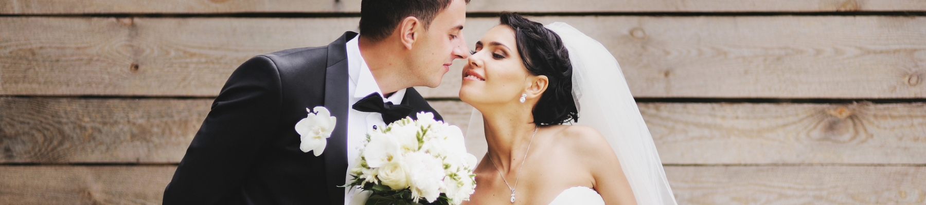 A bride and groom are dressed in wedding attire, leaning in for a kiss in front of a wooden backdrop, with the bride holding a bouquet.