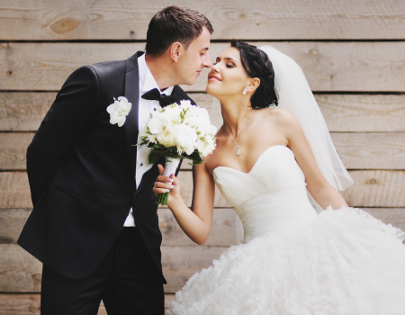 A bride and groom are dressed in wedding attire, leaning in for a kiss in front of a wooden backdrop, with the bride holding a bouquet.
