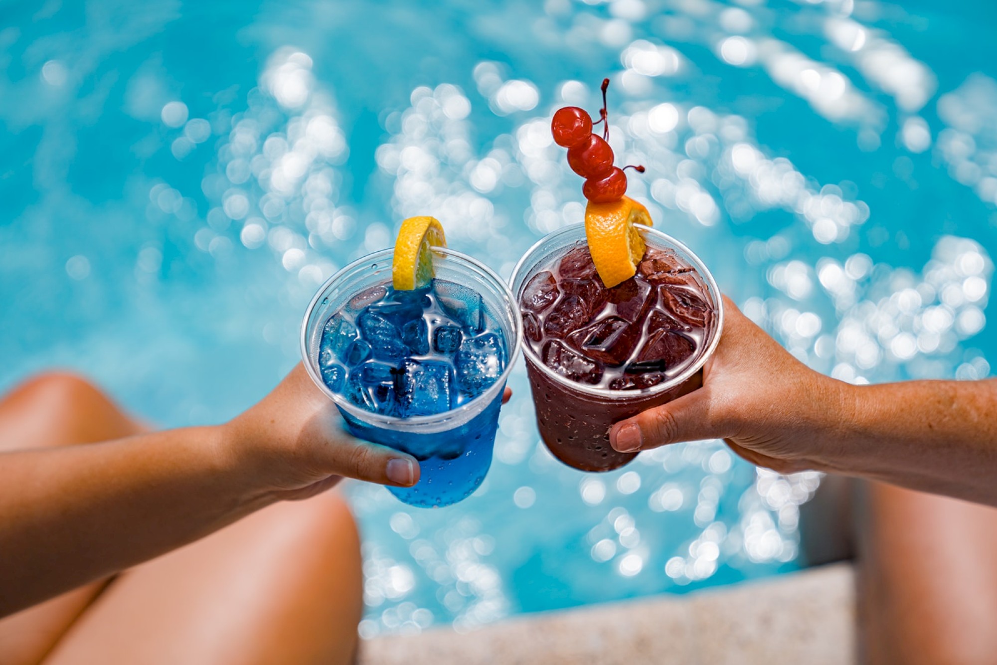 Two hands are holding colorful drinks, one blue with a lemon slice and one red with fruit garnishes, beside a pool.