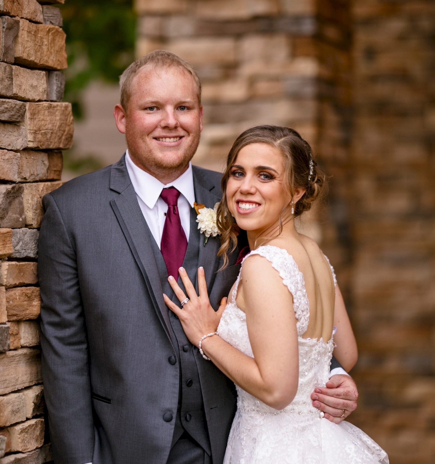 A smiling couple is standing outside, dressed in wedding attire. They are posing for a photo against a stone wall.