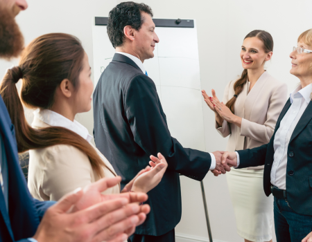 A group of people, dressed in professional attire, are in a meeting room with a flip chart; two people are shaking hands while others are clapping.
