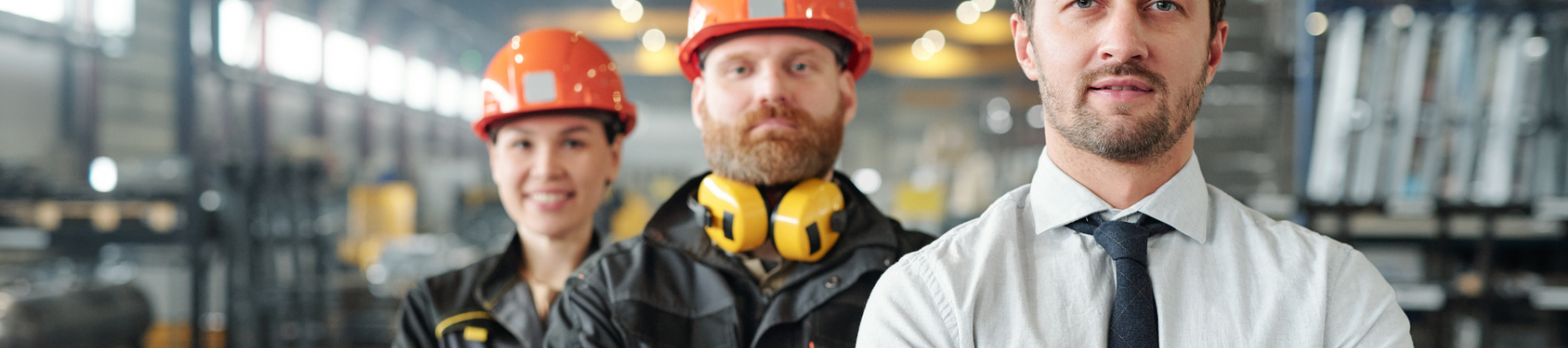 Three factory workers wearing safety equipment are standing in an industrial setting. The man in front is wearing a white hard hat and tie.