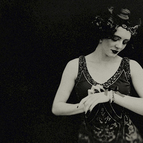 The image shows a person dressed in a vintage style, possibly from the early 20th century, looking down while adjusting something on their wrist.