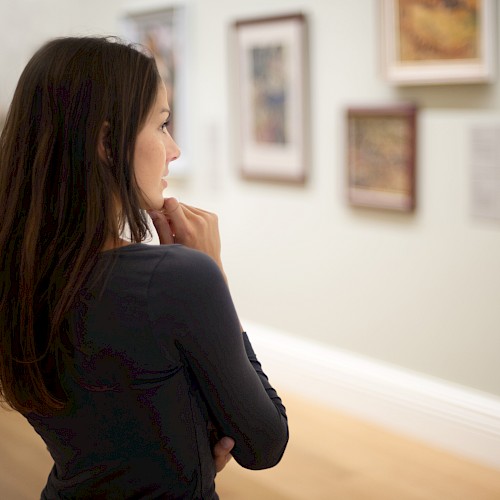 A person with long dark hair is thoughtfully observing framed artwork on the wall in what appears to be an art gallery.