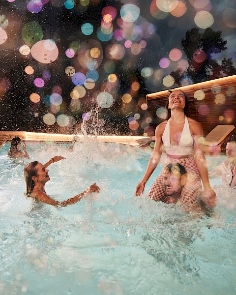 People are enjoying a night pool party with colorful bokeh lights in the background, and some are splashing and playing in the water.