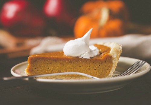 A slice of pumpkin pie topped with whipped cream on a plate, accompanied by a fork, with a blurred background including pumpkins.