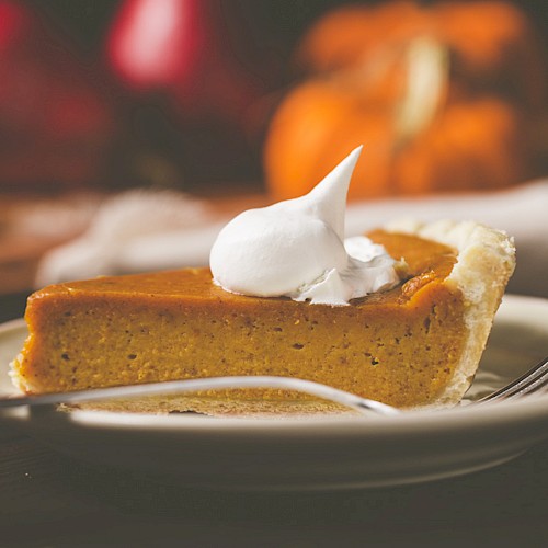 A slice of pumpkin pie topped with whipped cream on a plate, accompanied by a fork, with a blurred background including pumpkins.