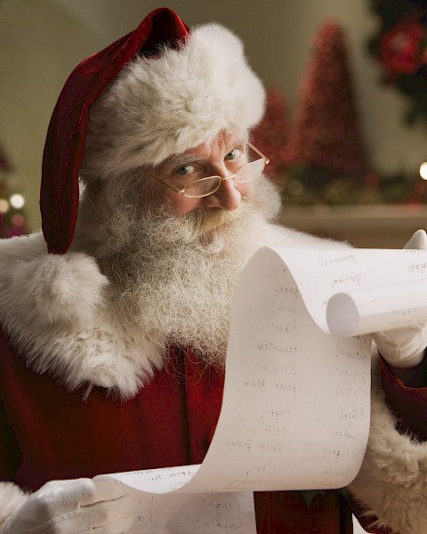 A person dressed as Santa Claus is holding a long list, with a decorated Christmas tree and festive background visible.