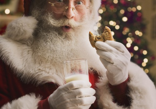 A person dressed as Santa Claus, holding a glass of milk and a cookie, with a decorated Christmas tree in the background.