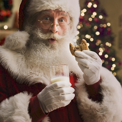 A person dressed as Santa Claus, holding a glass of milk and a cookie, with a decorated Christmas tree in the background.