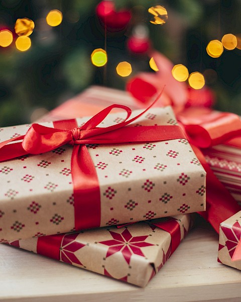 The image shows several wrapped Christmas gifts with red ribbons on a table, in front of a decorated Christmas tree with lights.