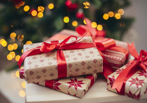 The image shows several wrapped Christmas gifts with red ribbons on a table, in front of a decorated Christmas tree with lights.